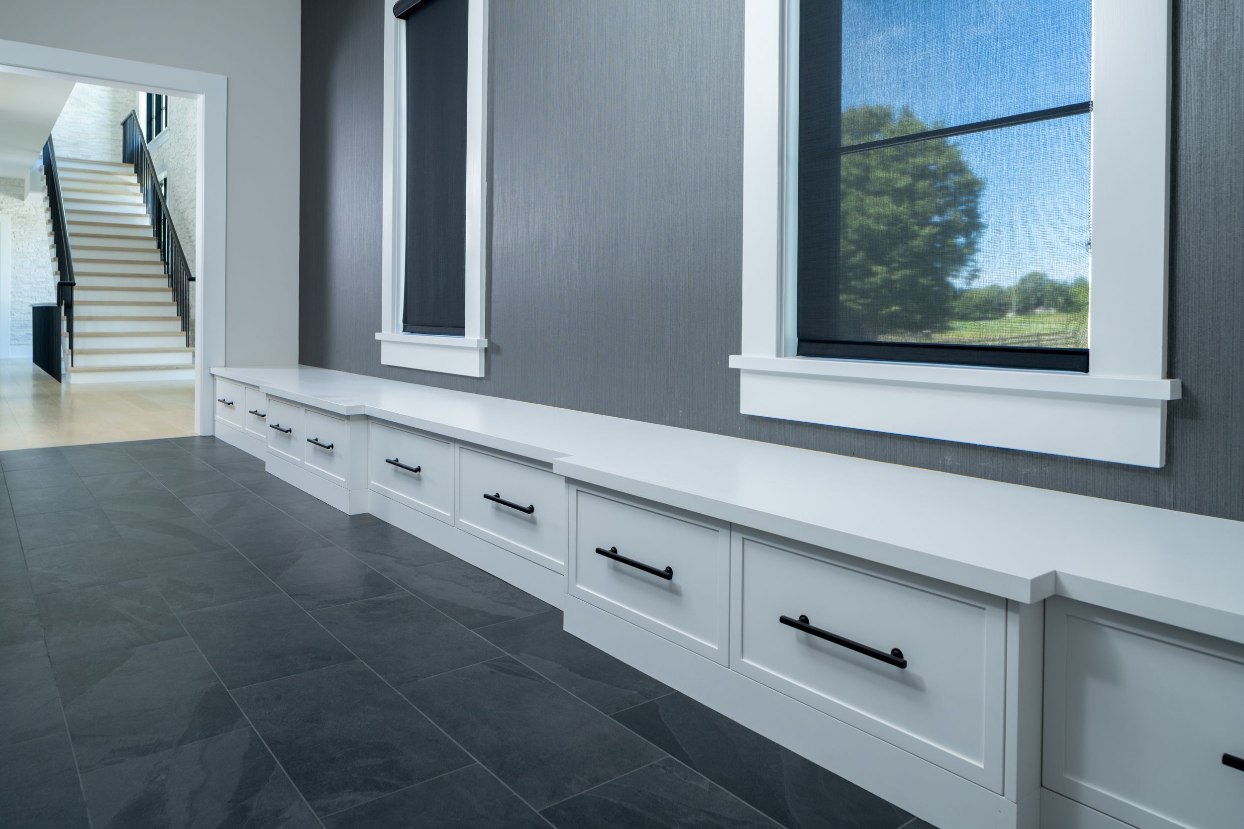 A modern entryway adorned with a row of streamlined white custom cabinets with black handles, set against a stylish gray wall, offering a view of the staircase, sourced from our custom cabinet gallery.