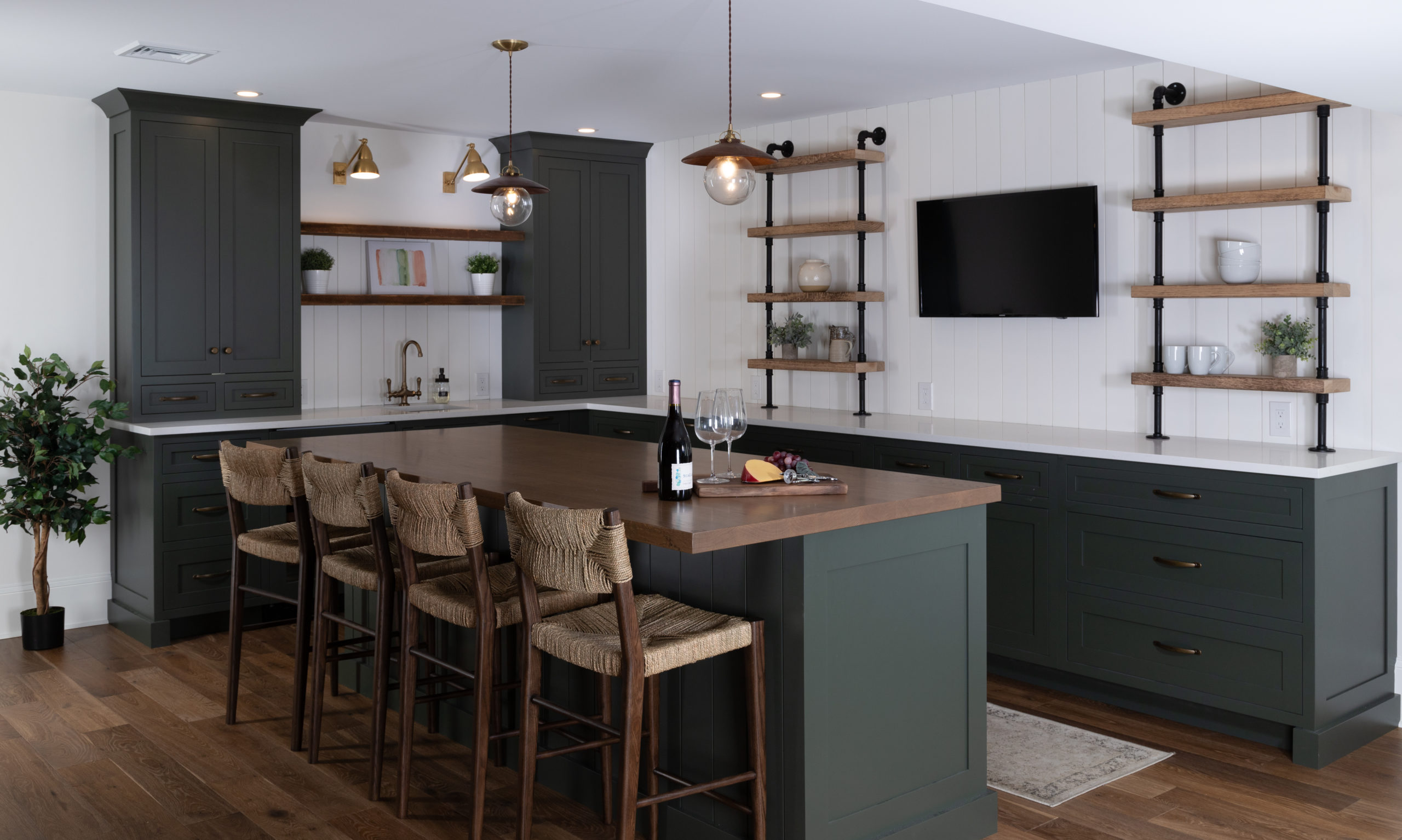 A modern home bar area featuring striking dark green custom cabinets, complemented by wooden countertops and open shelving, adorned with stylish pendant lighting.