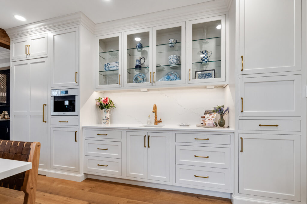 Traditional white luxury custom cabinetry with gold handles, elegant glass-front upper cabinets, and a marble backsplash, embodying timeless kitchen design.