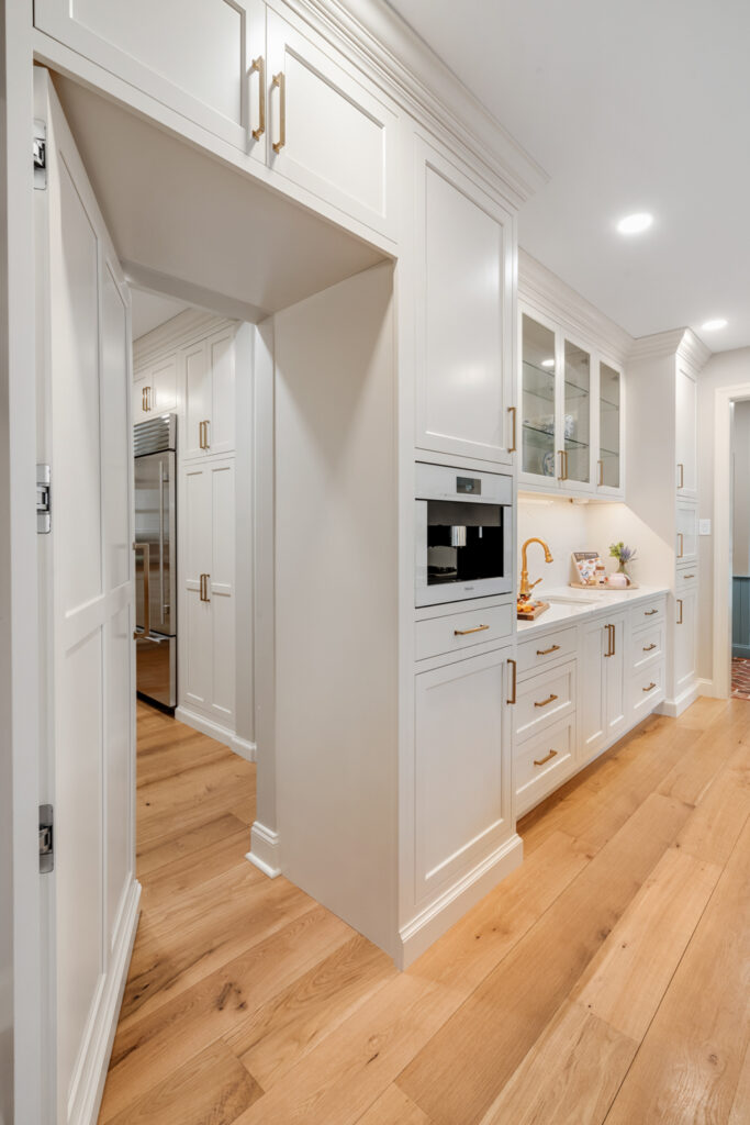 Open doorway in a kitchen revealing a custom-built luxury butler's pantry with matching white cabinetry and gold accents.